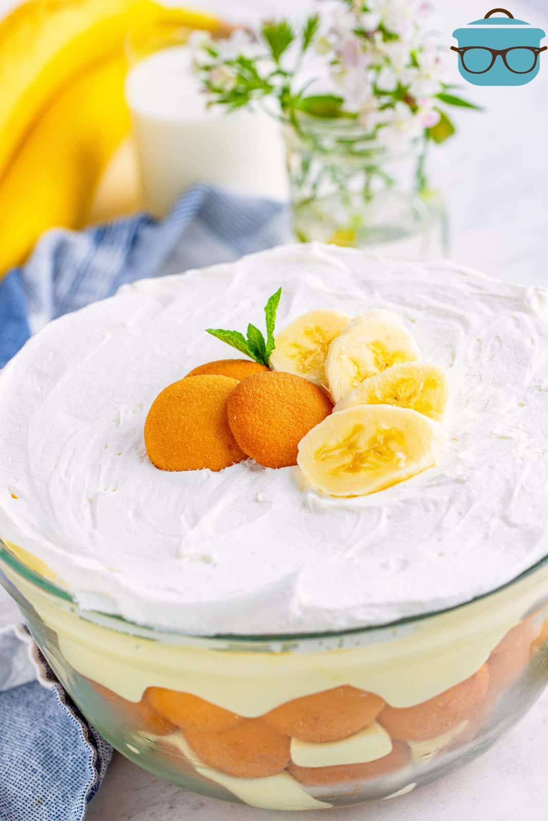 layers of banana pudding shown in a large glass serving bowl with a bunch of bananas in the background.